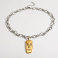 A silver chain necklace with a gold pendant shaped like a stylized face, featuring small blue stone eyes, photographed on a clean white background.