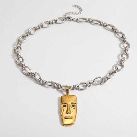A silver chain necklace with a gold pendant shaped like a stylized face, featuring small blue stone eyes, photographed on a clean white background.