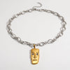 A silver chain necklace with a gold pendant shaped like a stylized face, featuring small blue stone eyes, photographed on a clean white background.