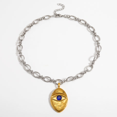 A silver chain necklace with a textured gold pendant shaped like a mummy wrapped around an eye, featuring a small blue gemstone in the center, displayed on a clean white background.