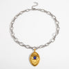 A silver chain necklace with a textured gold pendant shaped like a mummy wrapped around an eye, featuring a small blue gemstone in the center, displayed on a clean white background.
