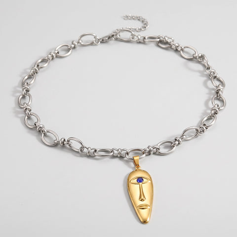 A silver-toned chain necklace with a gold-toned pendant shaped like a stylized face, featuring a small blue stone for an eye, displayed on a light gray background.