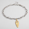 A silver-toned chain necklace with a gold-toned pendant shaped like a stylized face, featuring a small blue stone for an eye, displayed on a light gray background.