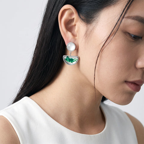 A close-up of a woman wearing a silver drop earring with a round white pearl-like top and a green semicircular dangling charm, paired with a white sleeveless top against a light gray background.