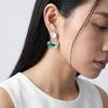 A close-up of a woman wearing a silver drop earring with a round white pearl-like top and a green semicircular dangling charm, paired with a white sleeveless top against a light gray background.