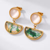 A pair of gold-tone drop earrings featuring round pale pink cabochon stones on the top and semi-circular clear resin pendants with green marbled swirls on the bottom, displayed on a clean white background.