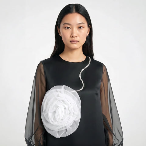 A model wearing a black long-sleeve top with sheer mesh sleeves and a large white fabric flower accessory attached near the chest, photographed against a light gray studio background.