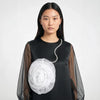A model wearing a black long-sleeve top with sheer mesh sleeves and a large white fabric flower accessory attached near the chest, photographed against a light gray studio background.