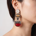 A close-up of a model's ear wearing a long, multi-tiered statement earring. The earring features a white enamel flower, a tan oval stone, a small black circular charm, and a large red teardrop-shaped stone, all set in silver-toned metal.