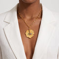A gold chain necklace with a textured flower pendant featuring small colorful gemstone accents, worn by a model in a white blazer.