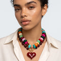 A close-up of a model wearing a chunky multi-strand statement necklace with colorful round beads and a red heart-shaped pendant, paired with a cream-colored collared shirt against a light gray background.