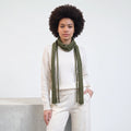 A model with an afro hairstyle wearing a cream-colored knit sweater, matching knit pants, and a knitted olive green scarf, standing against a white wall.