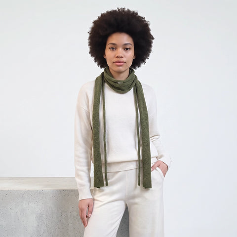 A model with an afro hairstyle wearing a cream-colored knit sweater, matching knit pants, and a knitted olive green scarf, standing against a white wall.