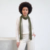 A model with an afro hairstyle wearing a cream-colored knit sweater, matching knit pants, and a knitted olive green scarf, standing against a white wall.