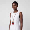 A model wearing a white sleeveless dress and a long brown cord necklace with stacked colorful wooden beads in tan, blue, and red, set against a plain light gray background.