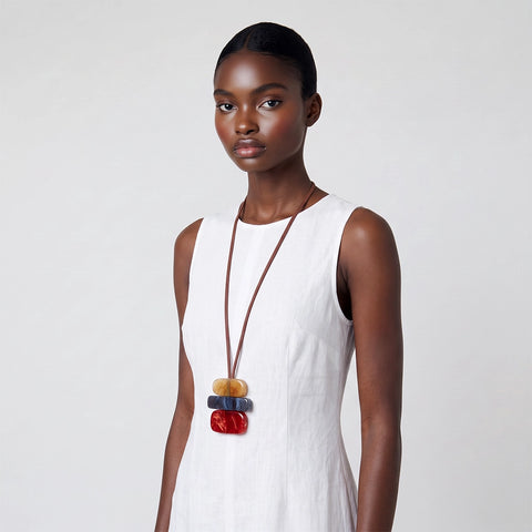 A model wearing a white sleeveless dress and a long brown cord necklace with stacked colorful wooden beads in tan, blue, and red, set against a plain light gray background.