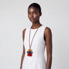 A model wearing a white sleeveless dress and a long brown cord necklace with stacked colorful wooden beads in tan, blue, and red, set against a plain light gray background.