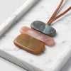Three polished stone pendants in brown, pink, and gray are arranged on a white marble tray with brown leather cords visible at the top.