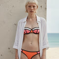 Woman wearing a colorful bikini with a white shirt over it, standing against a beach wall.