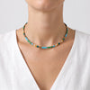 A close-up of a person's neck and upper chest wearing a thin beaded choker necklace. The necklace features small rectangular beads in turquoise blue, emerald green, and metallic gold, worn over a white V-neck top against a light gray background.