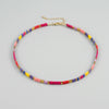 A beaded choker necklace with small rectangular beads in pink, yellow, purple, and red, accented with gold spacer beads and a gold chain extender, shown on a light gray background.
