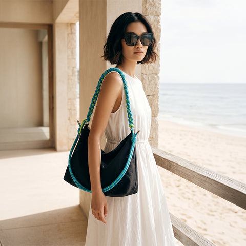 Woman in a white dress holding a black handbag with blue braided handle, standing by a window.