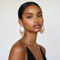 A woman with slicked-back black hair models a pair of large gold-tone hoop earrings adorned with cascading rows of small clear rhinestones, set against a plain light gray background.