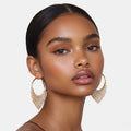 A close-up of a model wearing large gold hoop earrings with dangling beaded fringe, shown against a light gray studio background.