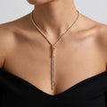 A gold Y-necklace with a drop chain, embellished with clear pavé stones, worn by a model in a black strapless top against a light gray background.