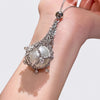 A silver-toned chain necklace with a large white pearl-like bead and intricate silver filigree, shown worn on a person's wrist against a light gray background.