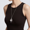 A silver-toned pendant necklace with a pearl and ornate filigree design, shown on a model wearing a dark brown ribbed tank top against a light gray background.