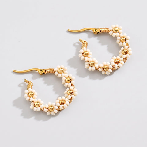 A pair of gold hoop earrings decorated with small white seed beads arranged in a daisy pattern, displayed on a light gray background.
