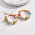 A pair of gold hoop earrings decorated with small multicolored flower beads, resting on a white marble surface.