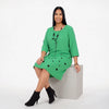 A woman wearing a bright green knee-length dress with black polka dots and a black beaded necklace, sitting on a light gray block against a white studio background.