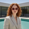 Woman with red hair wearing sunglasses and a beige blazer by a pool.