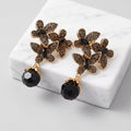 Pair of gold-tone floral drop earrings with dark crystal embellishments and faceted black bead drops displayed on a white marble block.