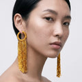 A close-up of a model wearing a pair of large gold-toned beaded dangle earrings with circular hoops and long fringe, shown against a plain light gray background.