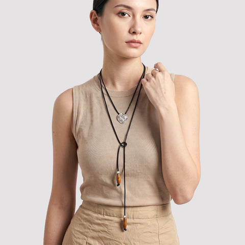 A woman wearing a beige ribbed sleeveless top and matching beige skirt, modeling a long black cord lariat necklace with a silver-toned circular pendant and two brown wooden beads, against a light gray studio background.