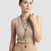 A woman wearing a beige ribbed sleeveless top and matching beige skirt, modeling a long black cord lariat necklace with a silver-toned circular pendant and two brown wooden beads, against a light gray studio background.