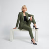 A woman with a short blonde bob sits on a white bench in a studio setting, wearing an olive green tailored blazer and matching trousers over a black top, with black high heels.