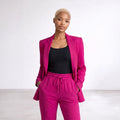 A model wearing a vibrant magenta pink two-piece suit consisting of a blazer and matching drawstring trousers, styled over a black tank top, set against a clean white studio background.