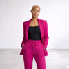A model wearing a bright magenta pink two-piece pantsuit consisting of a blazer and matching drawstring trousers, styled with a black tank top, against a clean white studio background.