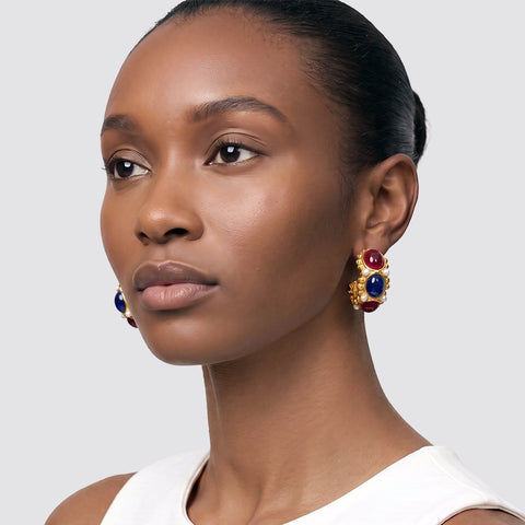 A close-up portrait of a woman wearing gold drop earrings with red and blue gemstone accents, set against a plain light gray background.