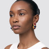 A close-up portrait of a woman wearing gold drop earrings with red and blue gemstone accents, set against a plain light gray background.