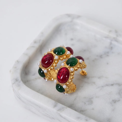A pair of gold-toned statement earrings with oval red and green cabochon stones and small pearl-like accents, displayed on a white marble tray against a light background.