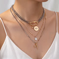 A close-up of a model wearing a layered necklace set. The set includes a chunky silver-toned chain, a gold-toned chain with a toggle clasp and lip charm, a delicate silver chain with a letter 'M' pendant, and a gold-toned chain with star and lightning bolt charms.