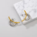 Two modern geometric hoop earrings with one polished gold curved bar and one polished silver hoop, displayed on a white marble block against a light gray background.