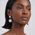 A close-up of a woman wearing double drop earrings with oval white cabochon stones in silver-toned settings, shown against a light gray background.