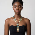 A model wearing a chunky gold statement necklace with large oval and round stones in blue, cream, and red, paired with a black strapless top against a light gray background.