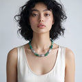 A model with short, curly black hair is wearing a silver and green beaded necklace with a cream-colored ribbed tank top, photographed against a neutral light gray background.
